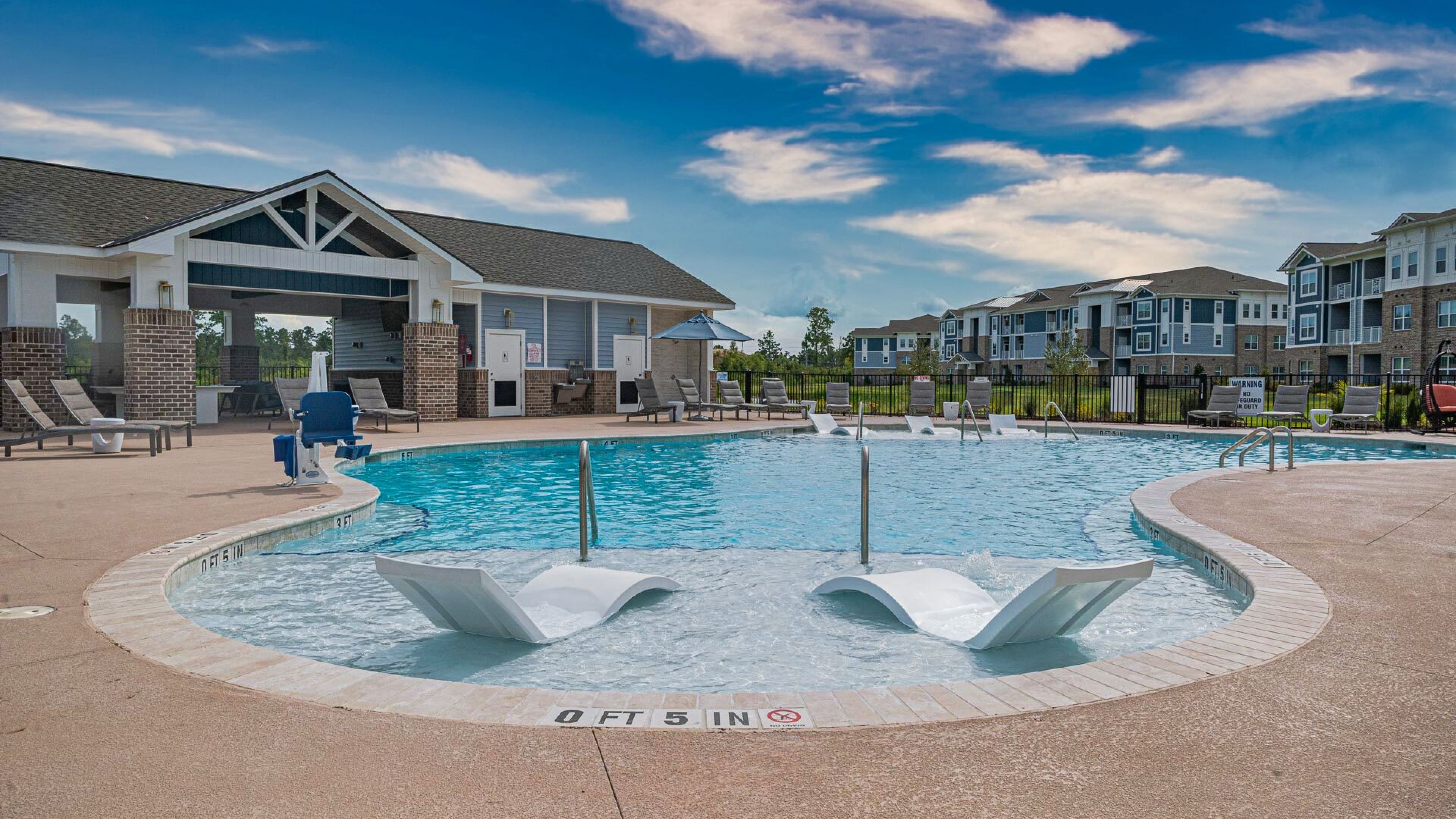 Resort-style community pool flanked by sun loungers and a covered seating area, with a backdrop of apartment buildings.